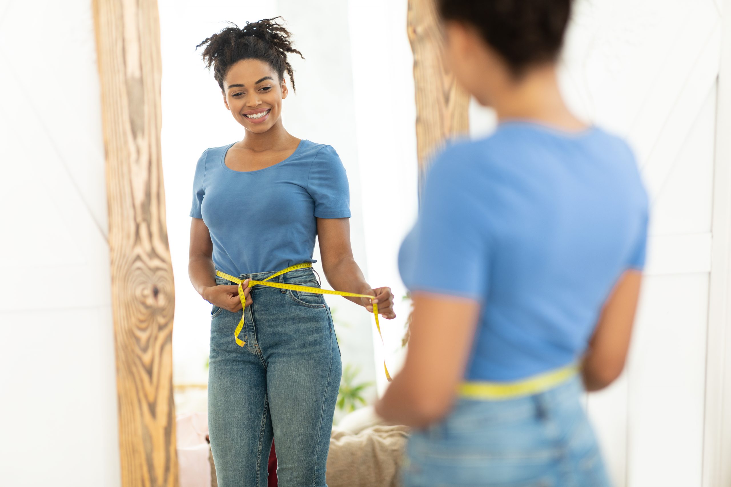 Happy Black Girl After Weight Loss Measuring Thin Waist With Tape Standing Near Mirror At Home. Slimming Concept. Selective Focus
