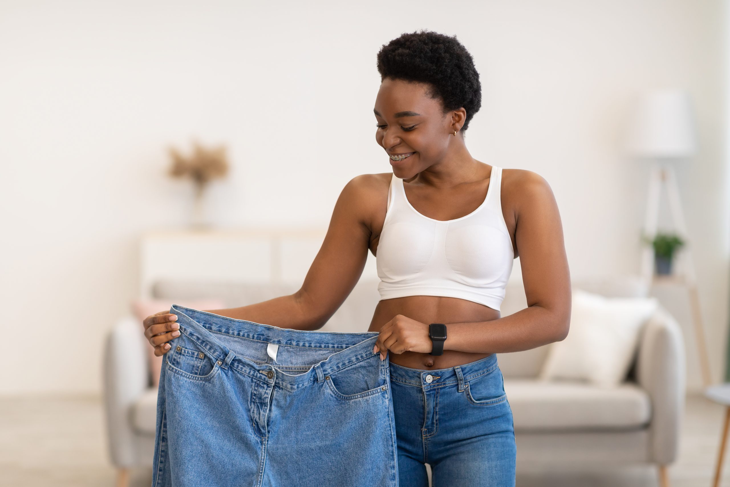 Joy Of Weight Loss. Black Lady Holding Oversized Jeans Too Big After Great Weight-Loss And Successful Slimming Standing Posing At Home. Dieting Motivation Concept